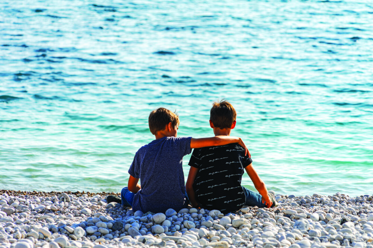 Two boys sitting on the beach, talking and throwing rocks into the sea.