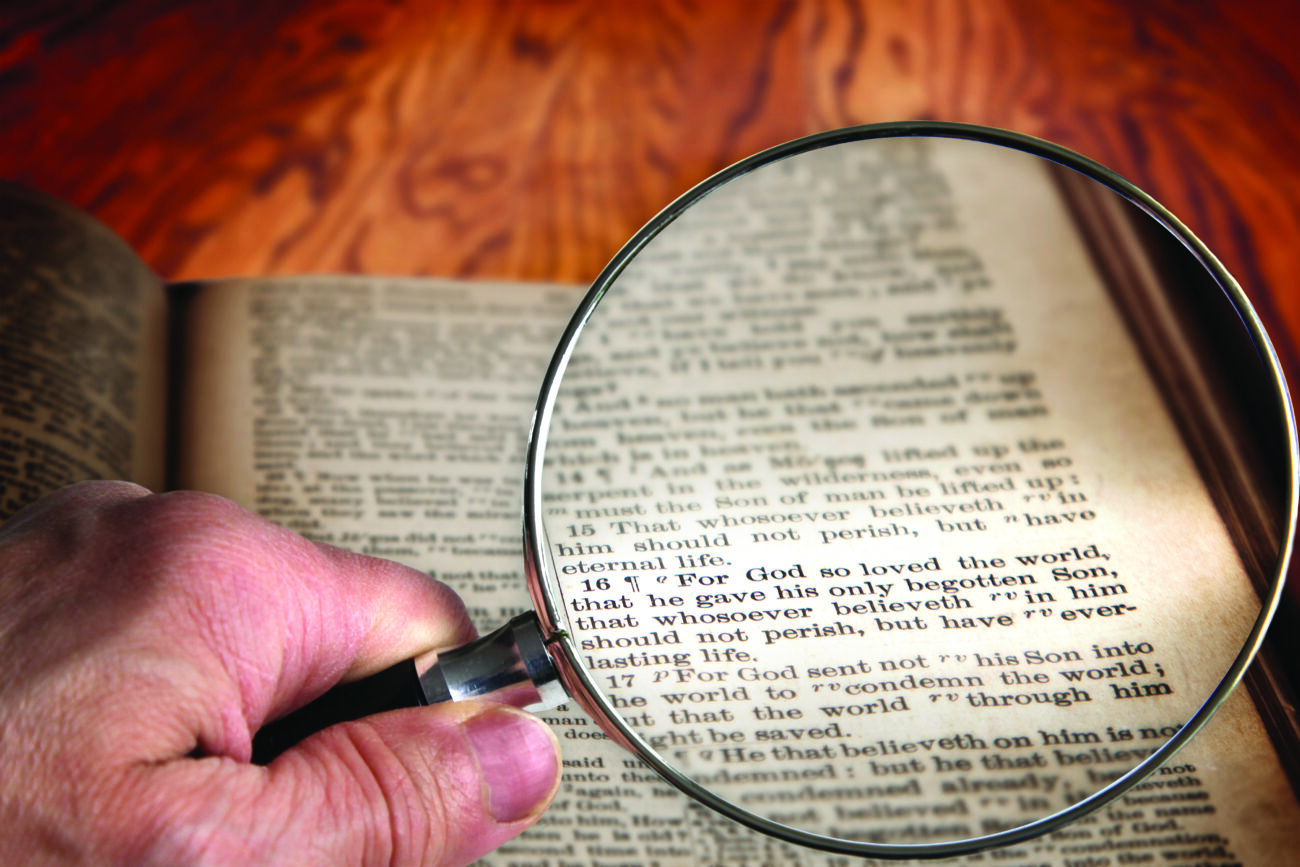 Hand holding magnifying glass over an old Bible opened to the famous verse of John 3:16. This translation is King James, which is public domain.